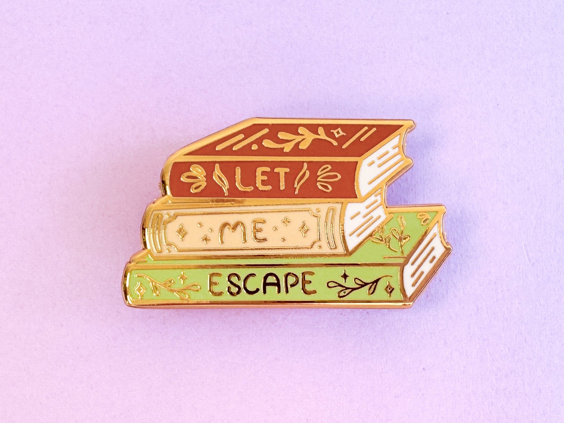Golden enamel pin of a stack of 3 books with decorative designs on them. The spines read "Let. Me. Escape."