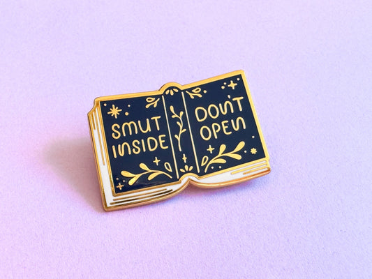 Golden enamel pin of a book laying down. The cover is dark blue and reads "Smut inside, don't open" in gold with leaves and stars decorating the cover.